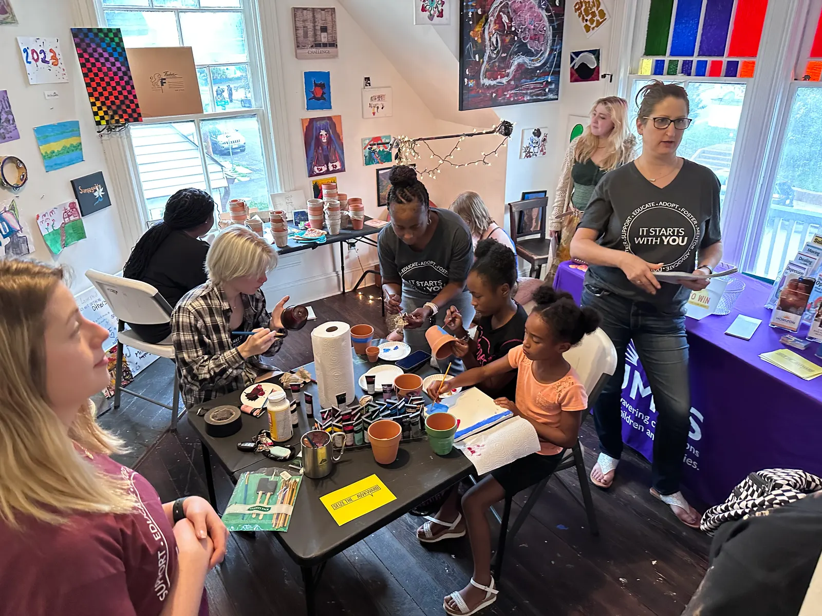 Neighbors of all ages gather around a table to make art together in a room lined with paintings
