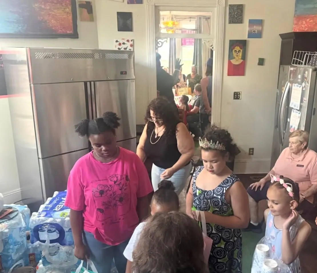 Kids and families in the kitchen at The Clarity House beside stacked cases of drinking water